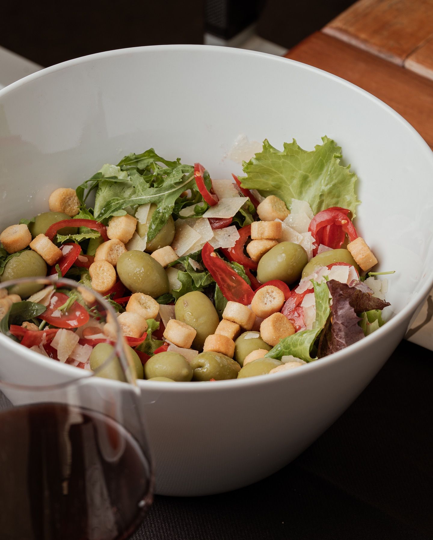 Insalata in una ciotola bianca con olive, pomodori, crostini e verdure; vicino a un bicchiere di vino.