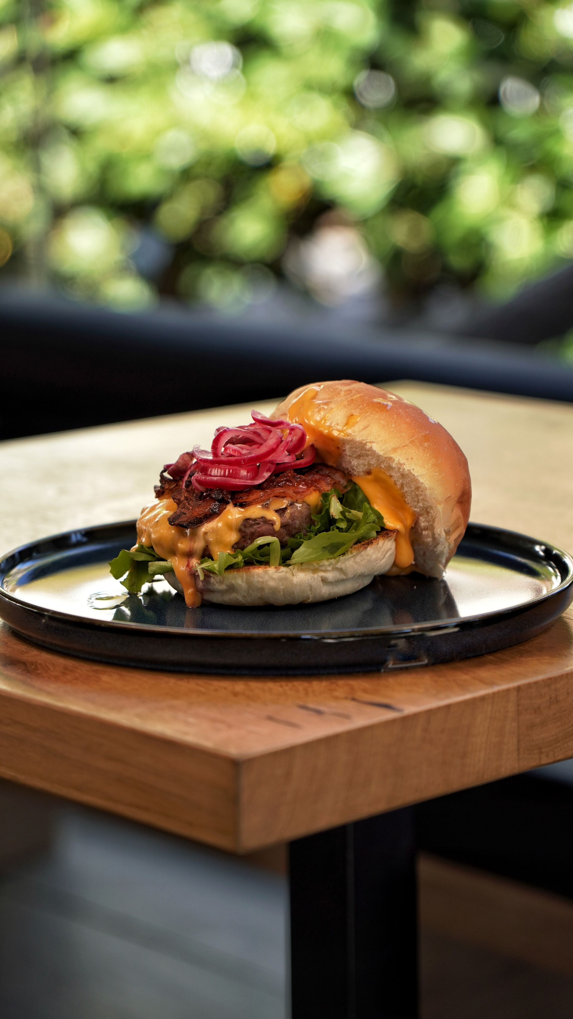 Hamburger su un piatto nero, con formaggio, verdure e un condimento. Tavolo di legno, sfondo verde sfocato.