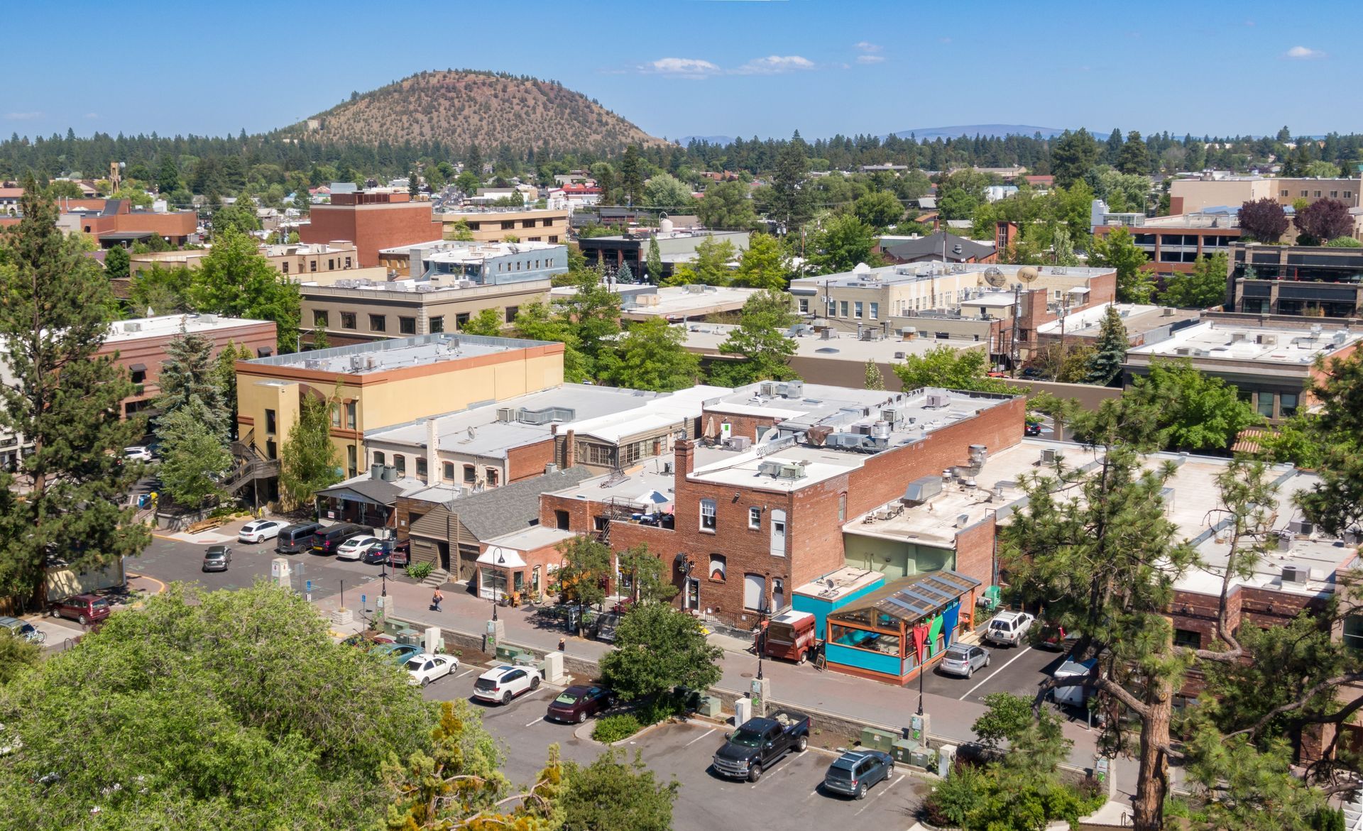 An aerial view of a downtown area in Bend, Oregon, featuring Pilot Butte in the background under a clear blue sky.