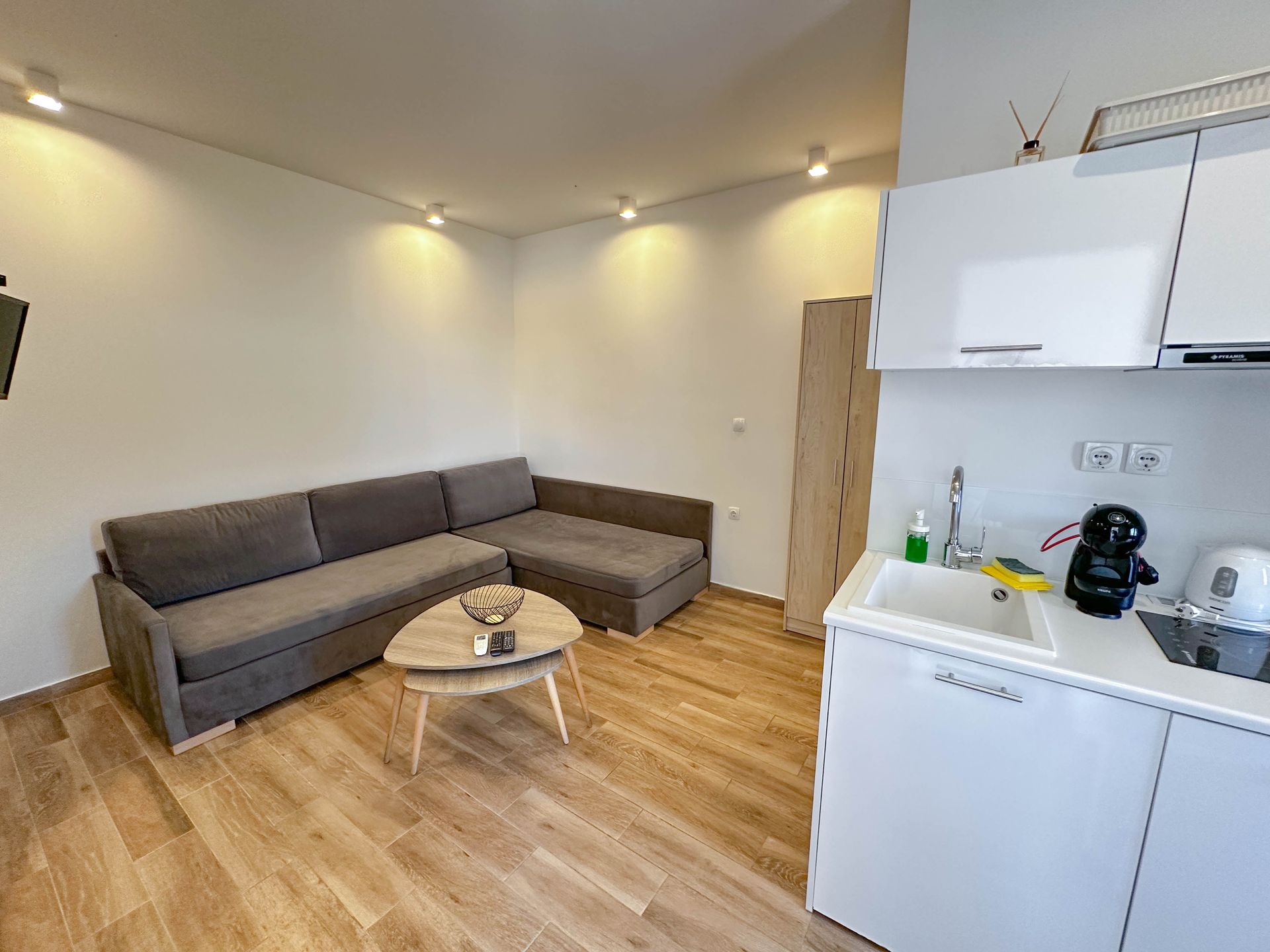 Small apartment interior with a gray sofa, kitchen, and wooden floor.
