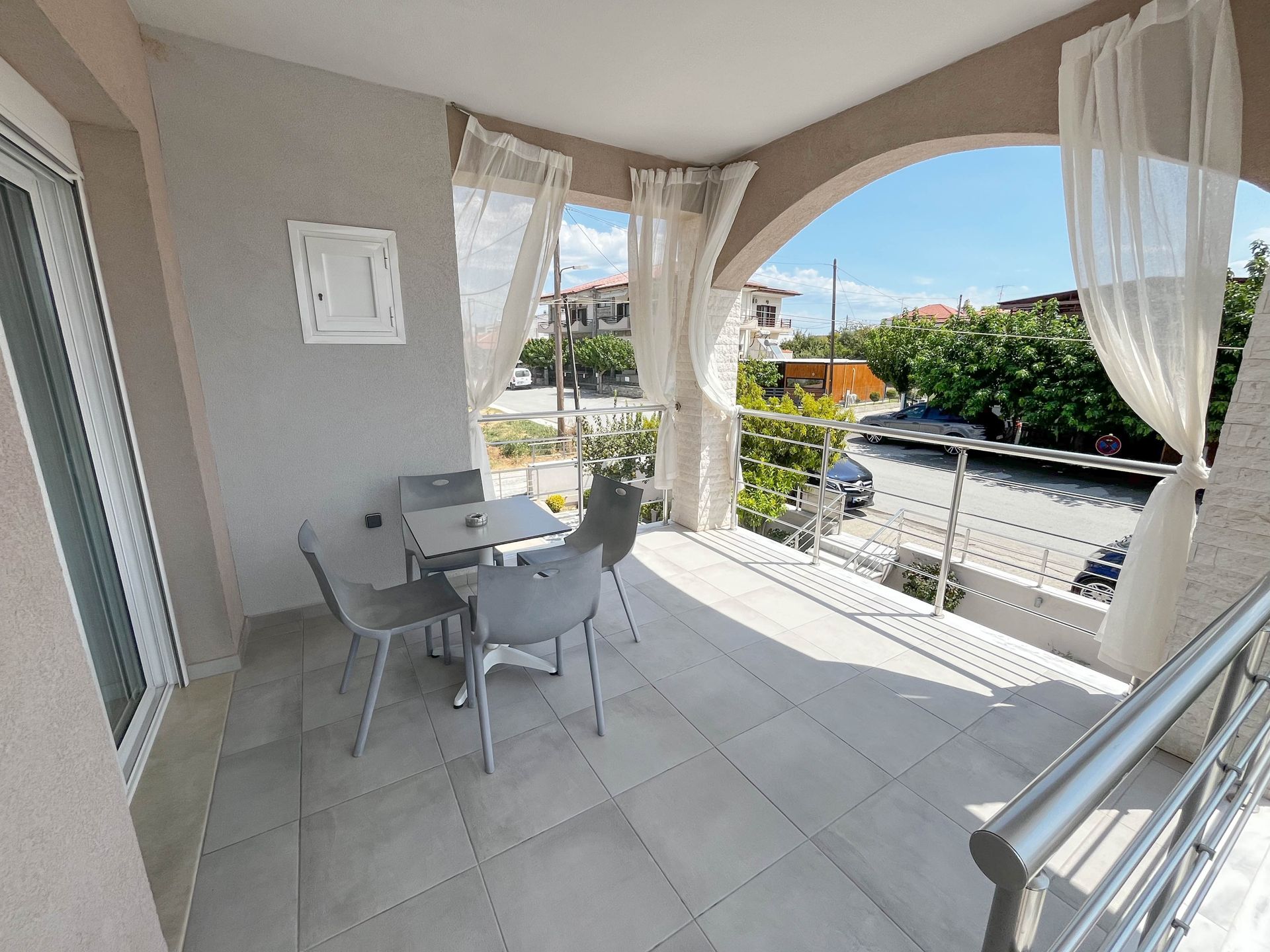Balcony with gray tile, table and chairs. Street view framed by arches and sheer curtains.