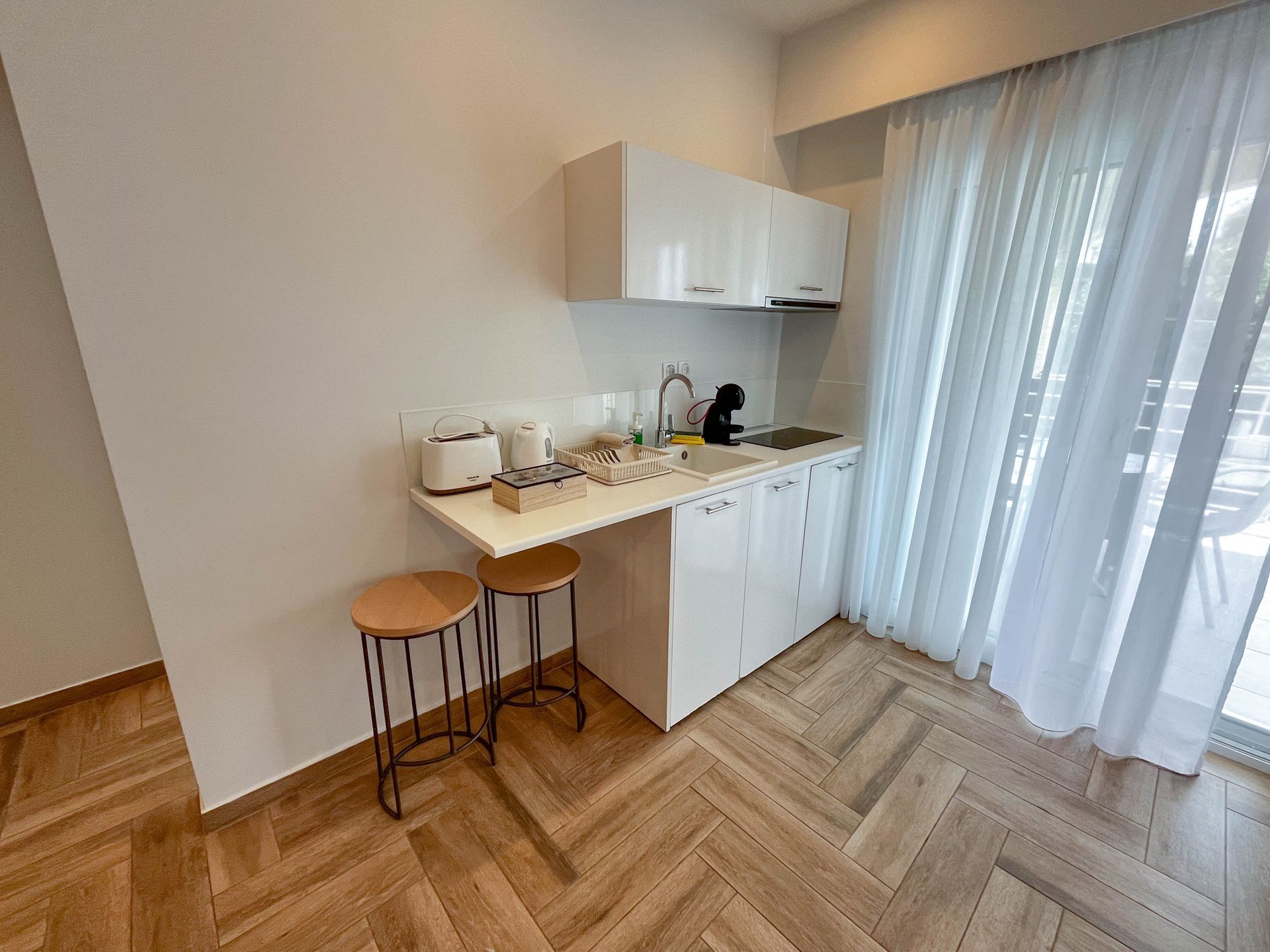 Small kitchen with white cabinets, breakfast bar, and two stools. Herringbone wood floors.