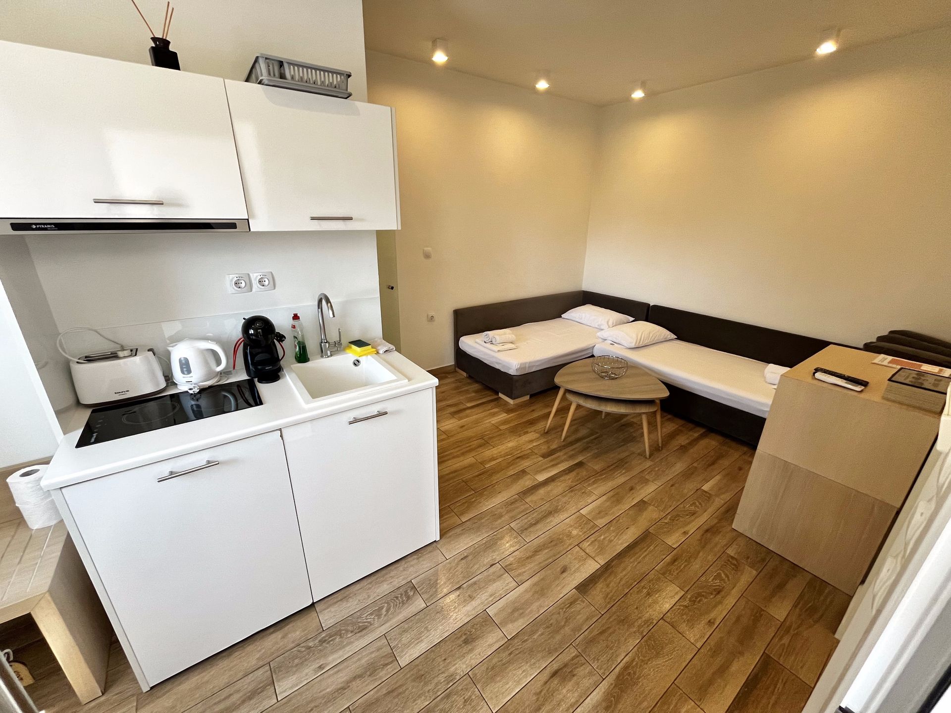 Small studio apartment with kitchenette, seating area, wooden floors, and white cabinetry.