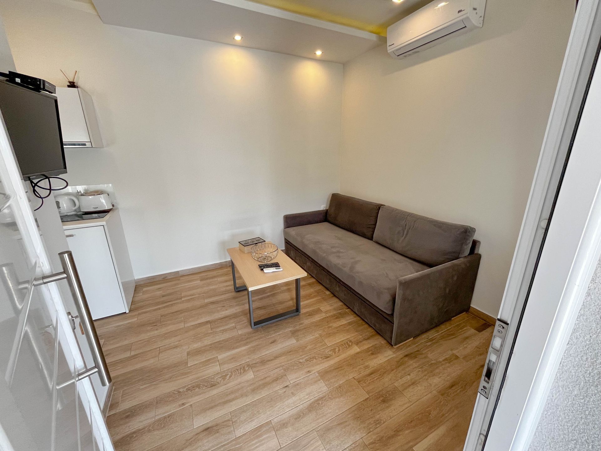 Small room with sofa, coffee table, TV, and mini-fridge. Wooden floor, white walls.