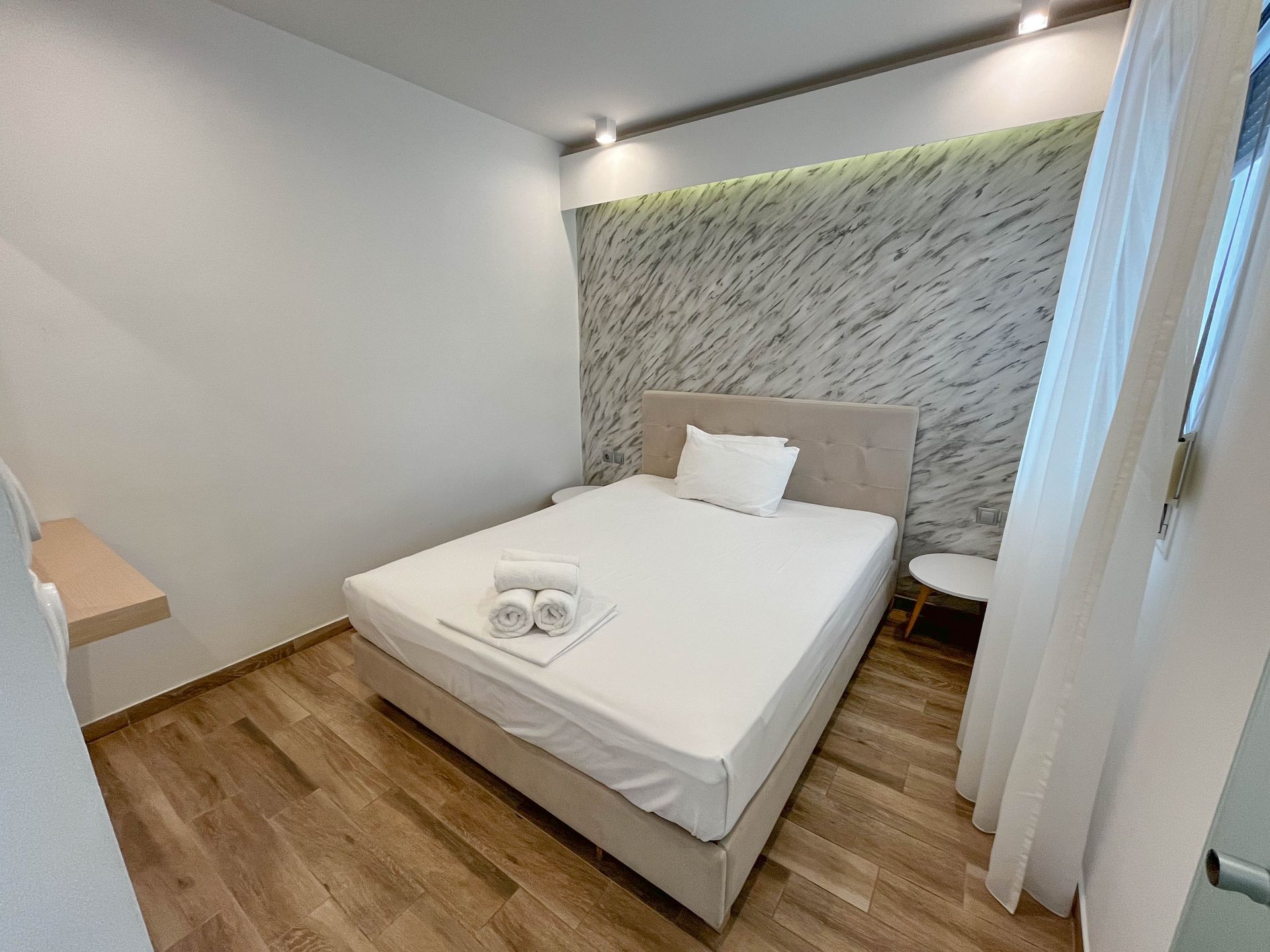 Bedroom with a bed, small side table, and textured wall behind the headboard. Wooden floors and white linens.
