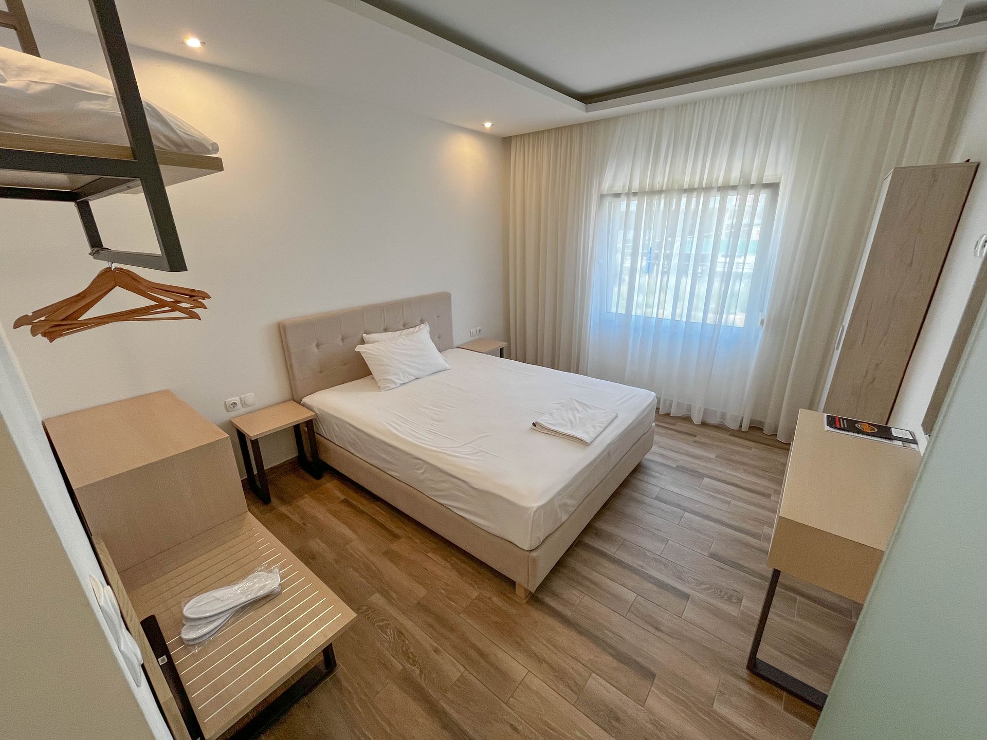 Bedroom with a bed, nightstand, dresser, and a window with sheer curtains. Wooden floors.