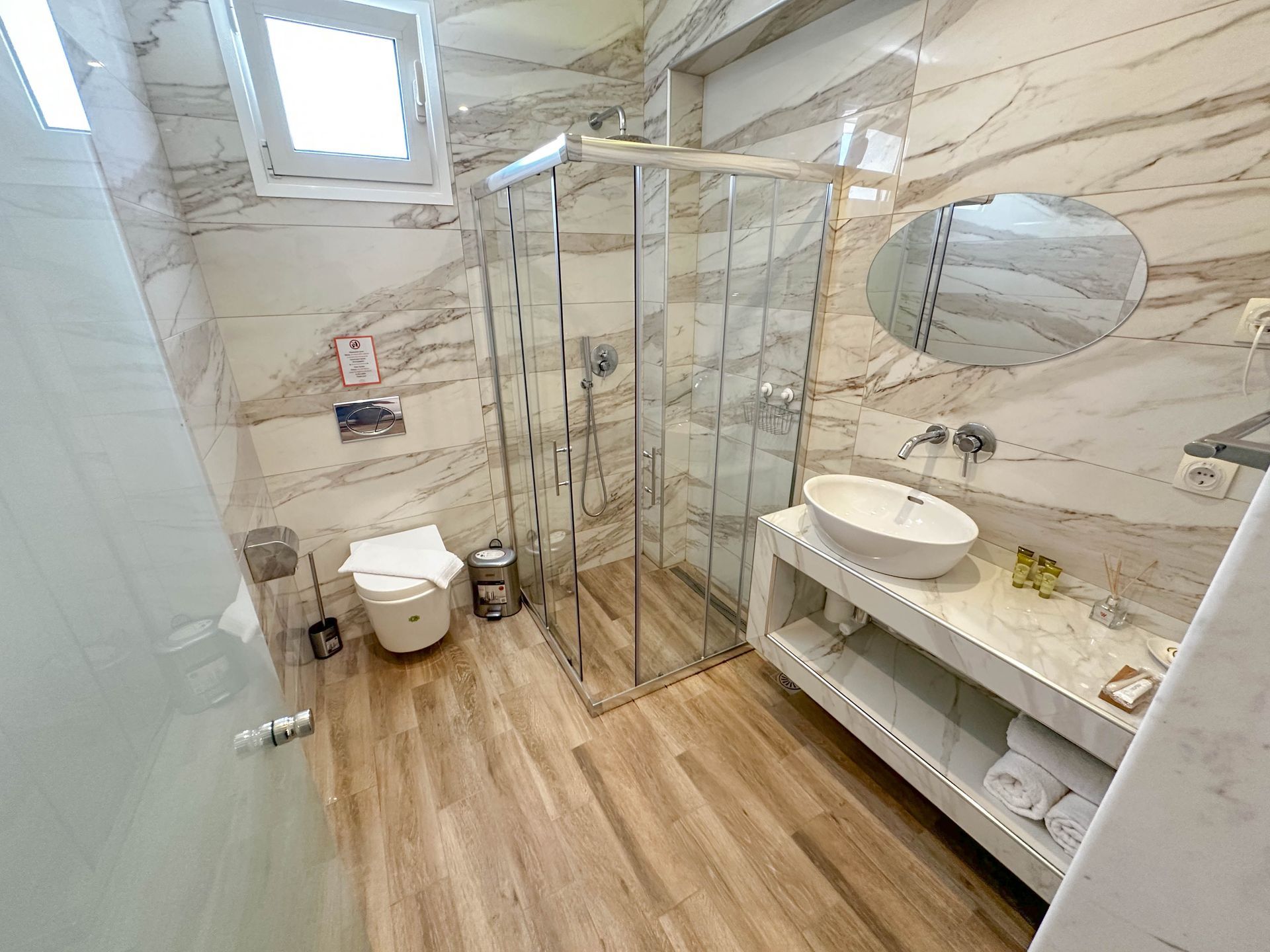 Modern bathroom with marble walls, glass shower, wooden floor, toilet, sink, and mirror.