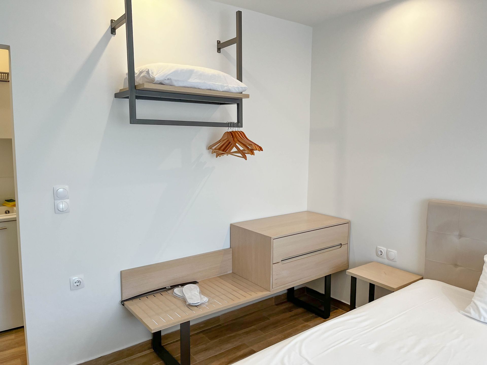 Bedroom interior with light-colored walls, bed, desk, storage unit, shelf, and wooden hanger.