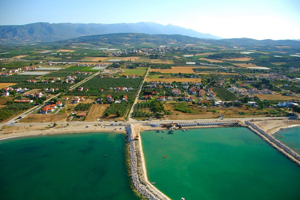 Aerial view of a coastal town with green water, beaches, breakwaters, and agricultural land with mountains in the background.