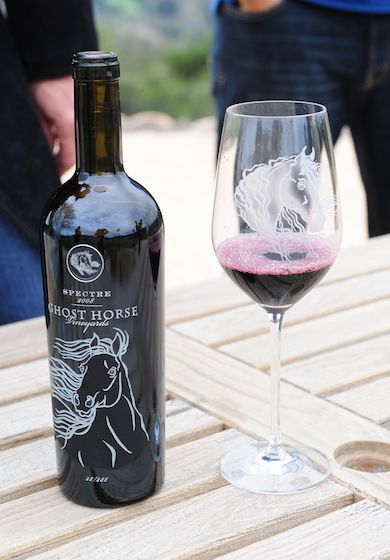 A bottle of Ghost Horse red wine and a matching glass etched with a horse image sitting on a light wooden table outdoors.