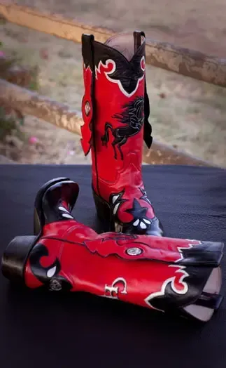 A pair of red and black cowboy boots featuring a horse emblem, set against a wooden fence background.