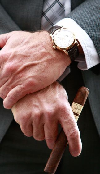 A person in a suit, wearing a watch, holding an unlit cigar in their crossed hands.