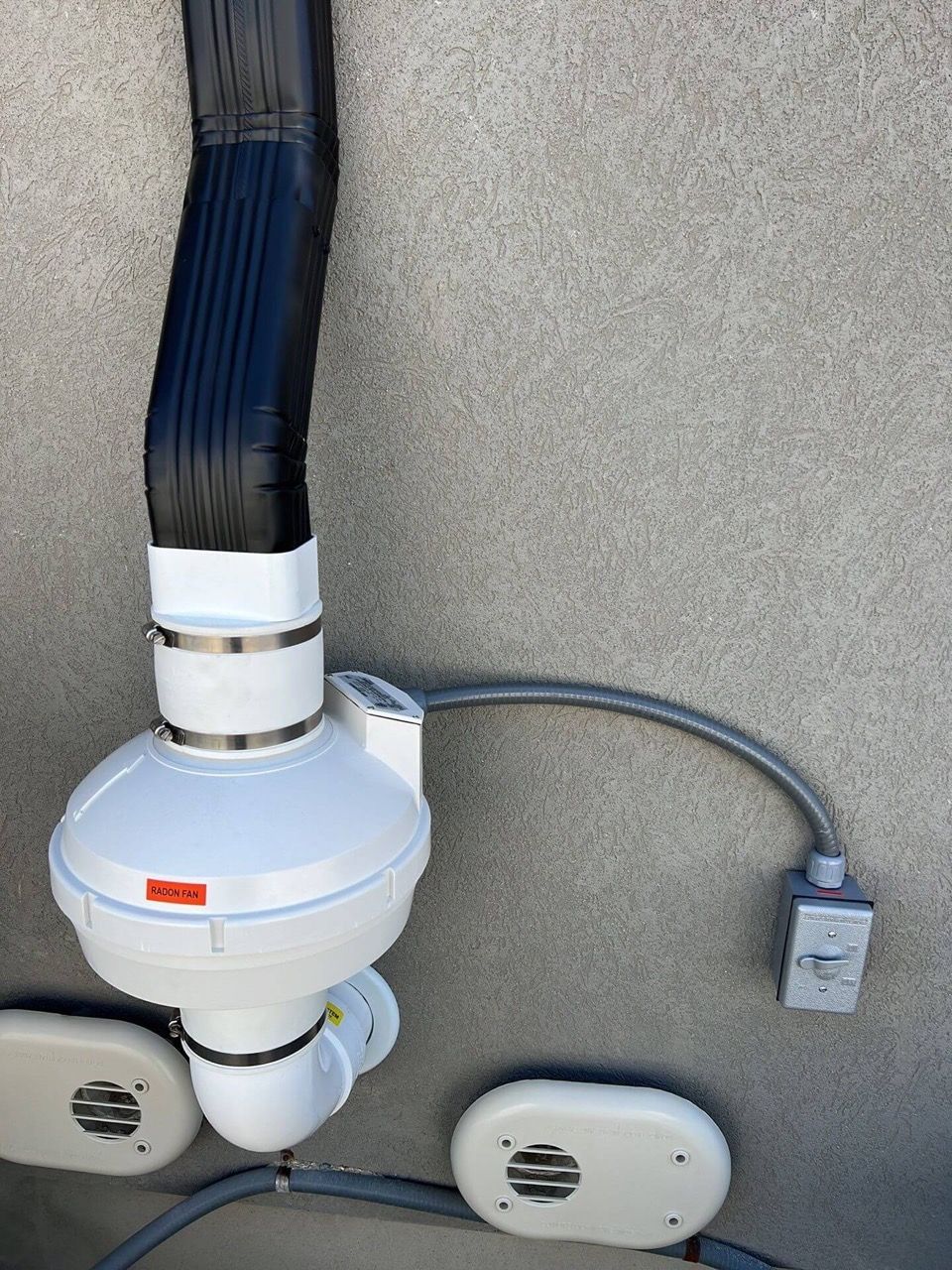 White ceiling exhaust fan with black ducting and a corded control mounted on a beige wall.