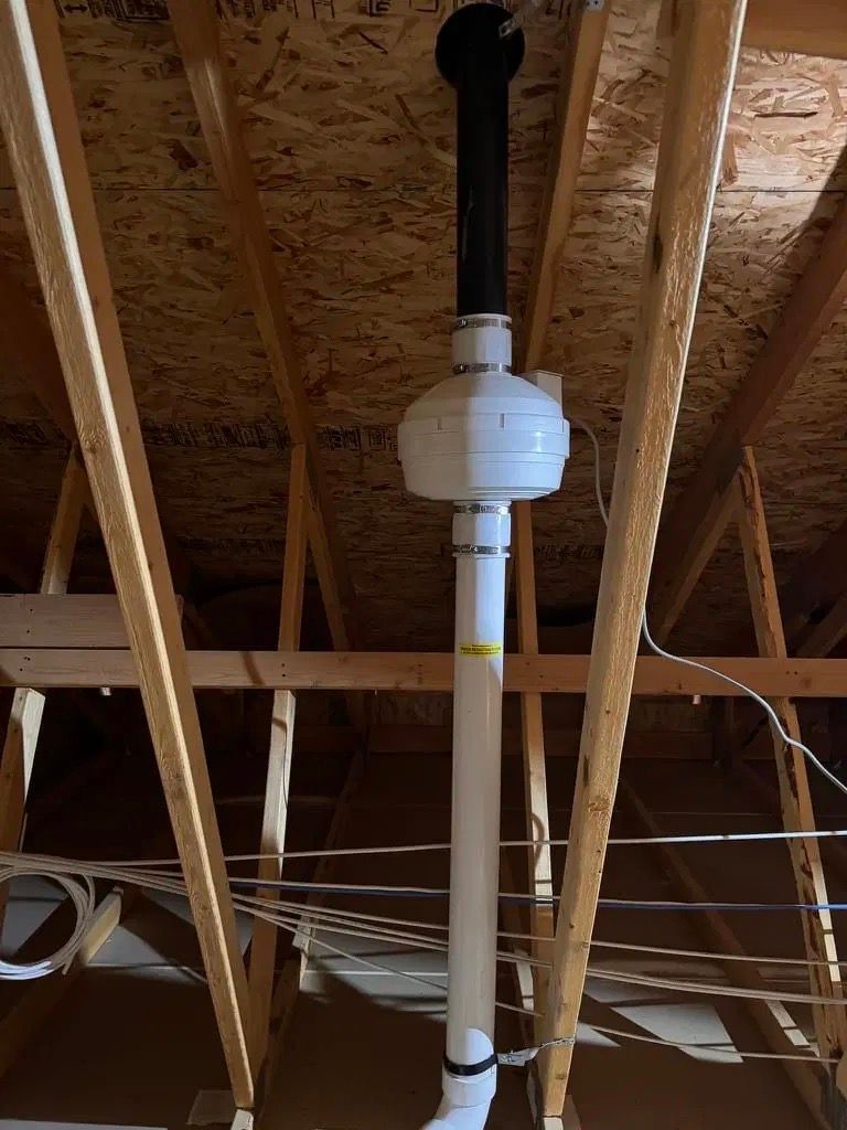 Vertical white vent pipe with a black top passing through unfinished attic rafters.