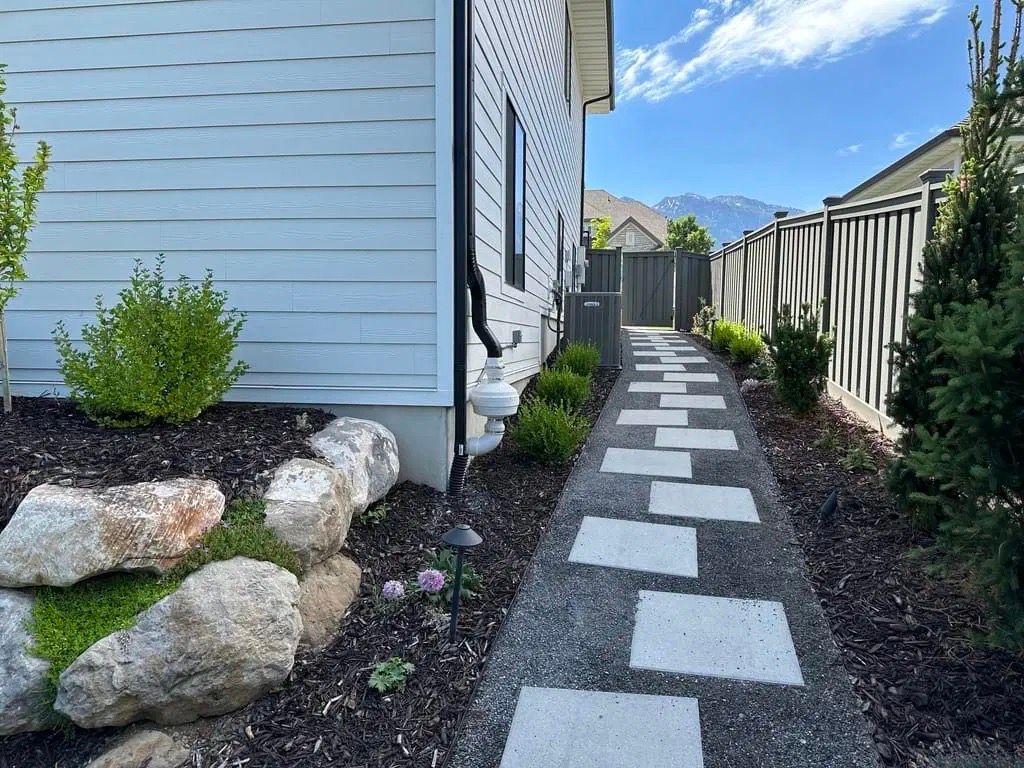 Narrow backyard path with stepping stones between a house and fence, bordered by rocks and shrubs.
