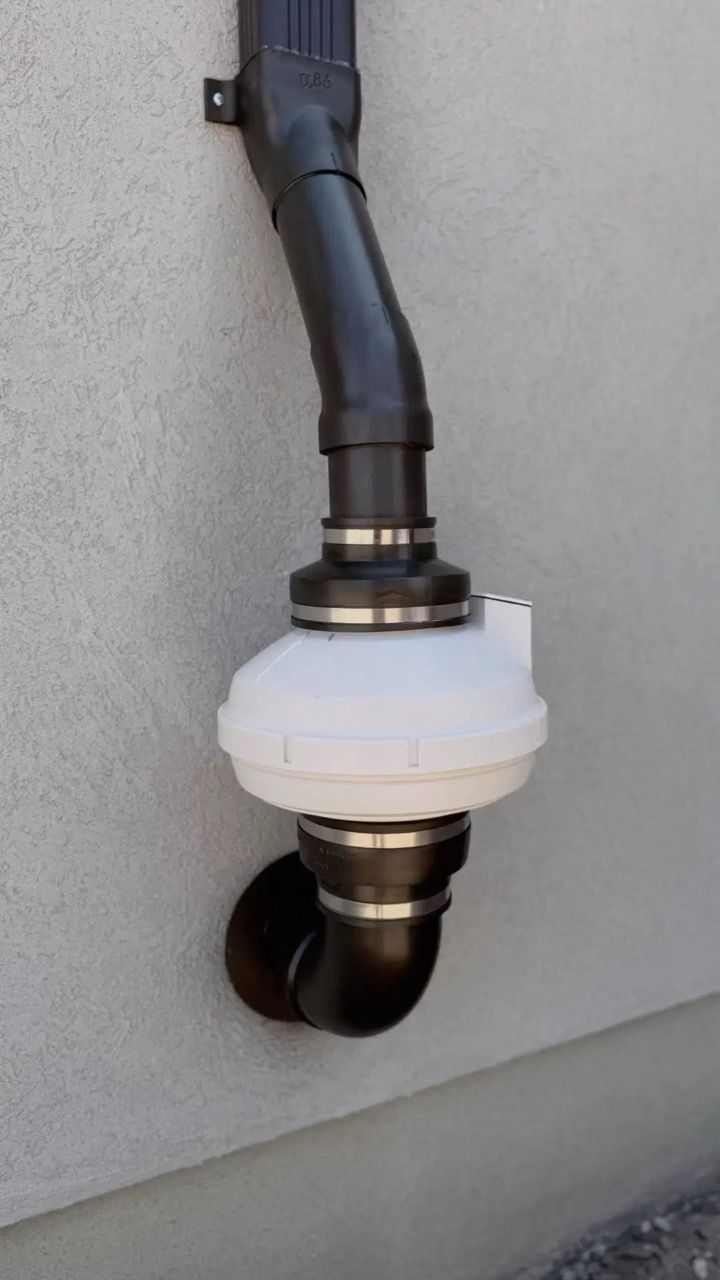 White plumbing vent/filter unit on black pipes mounted to a beige exterior wall