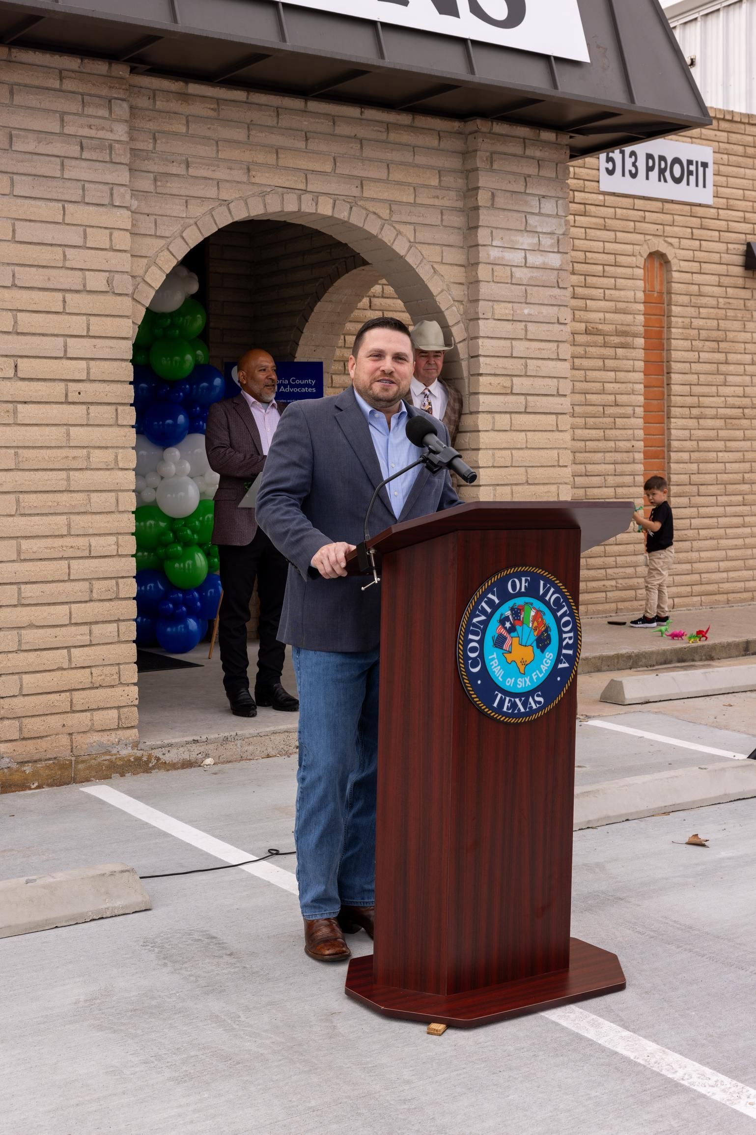 Man speaking at podium outside building; blue blazer, jeans, balloons.