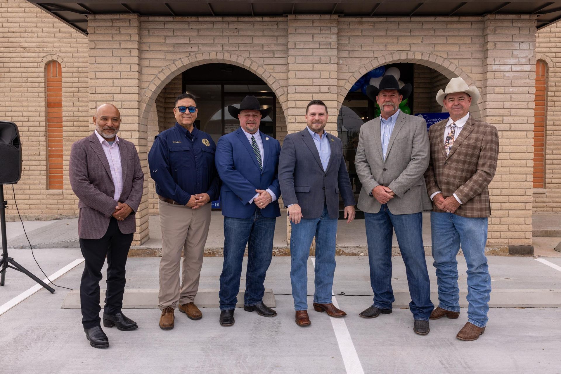 Group of six men posing in front of a brick building. Several wear cowboy hats and suits.
