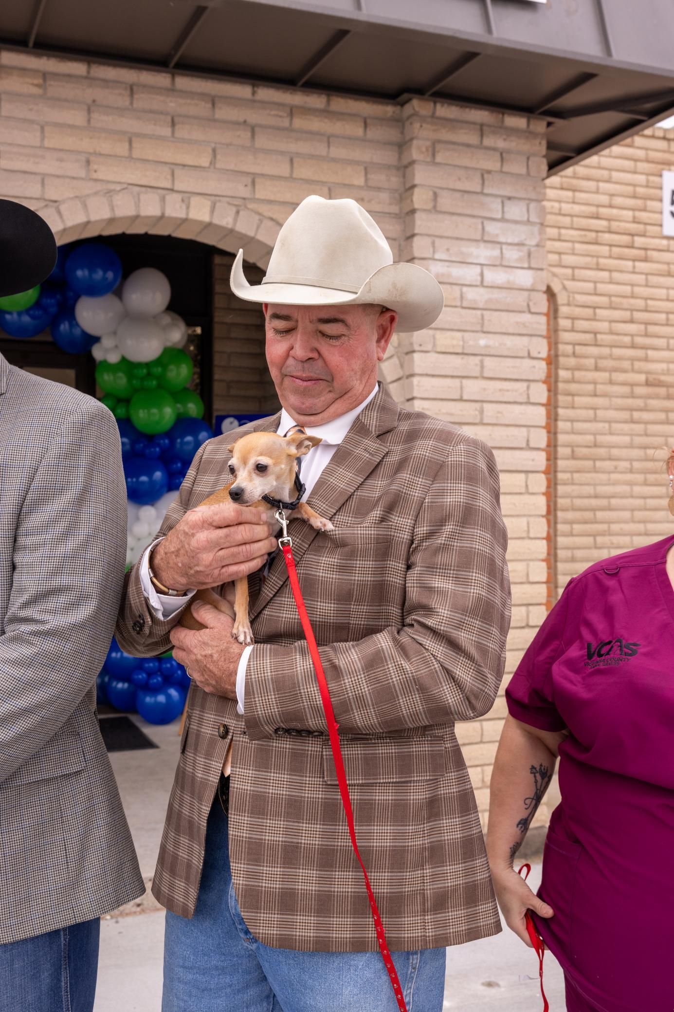 Man in cowboy hat and plaid blazer holds a small dog on a leash outside a building with balloons.