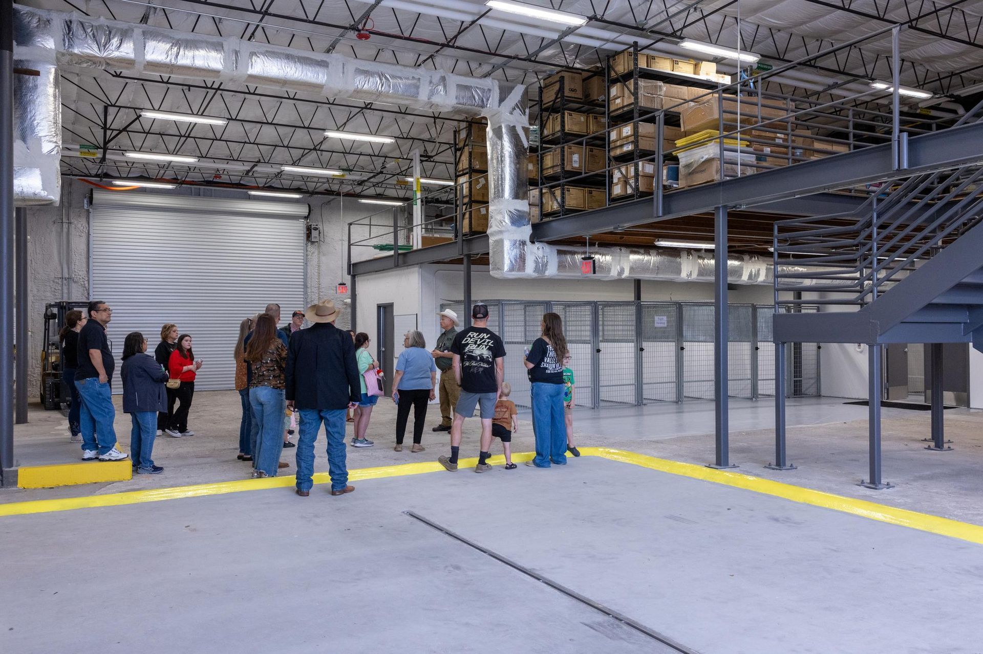 Group of people inside a warehouse; boxes stacked on a mezzanine level, metal structure, yellow and gray floor.