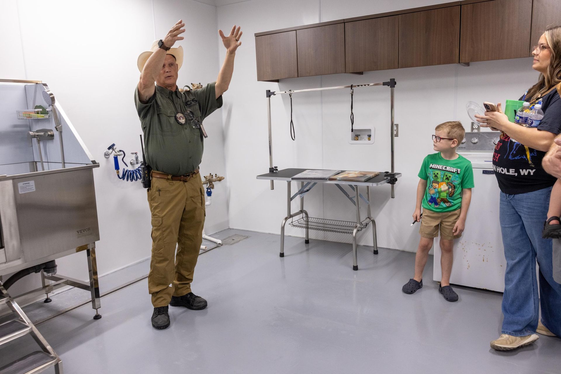 Man in uniform gestures, giving a tour inside a bright room with a grooming table and pet washing station.