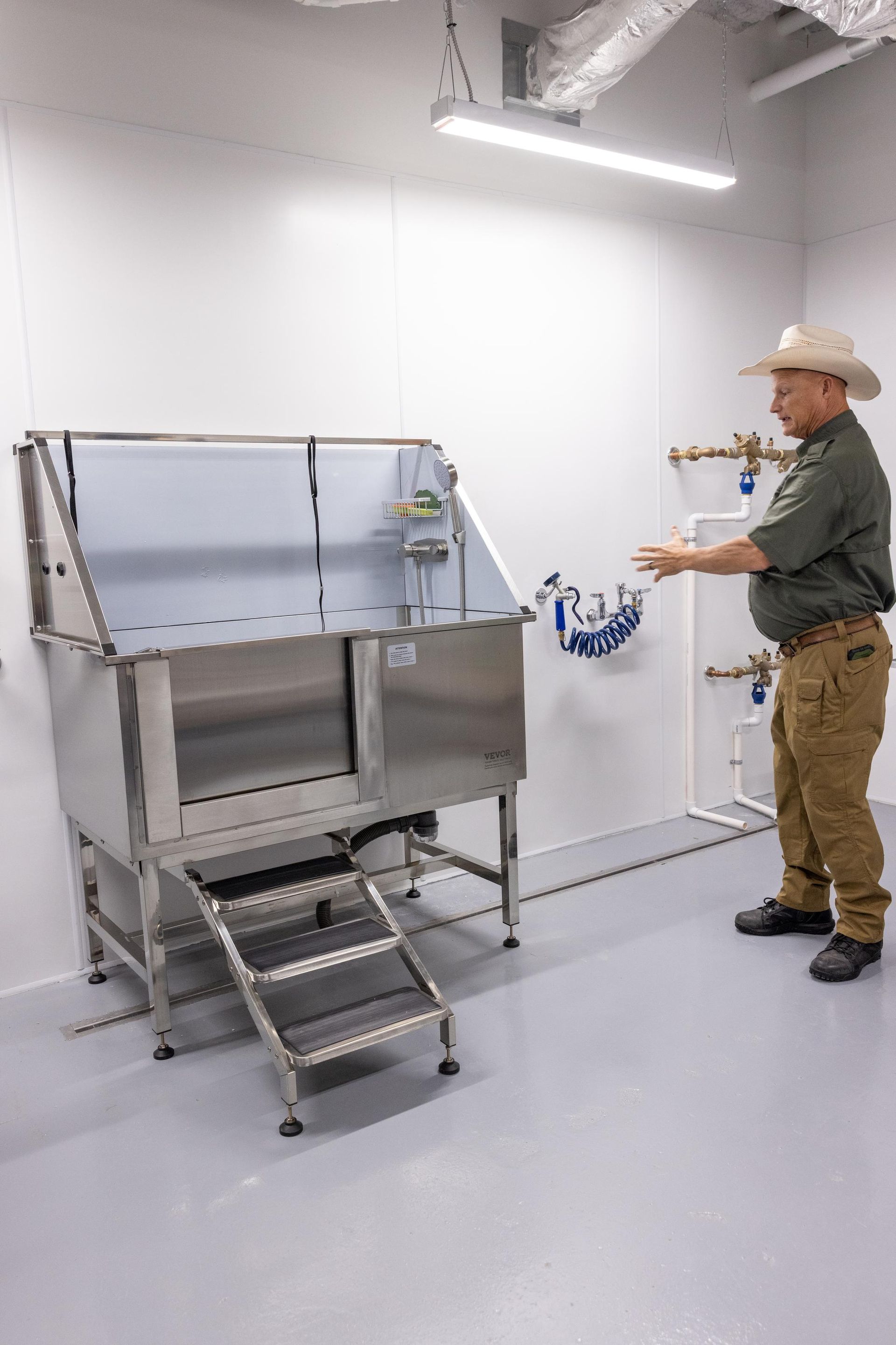 Man in a cowboy hat gesturing towards a stainless steel pet washing station in a white room.
