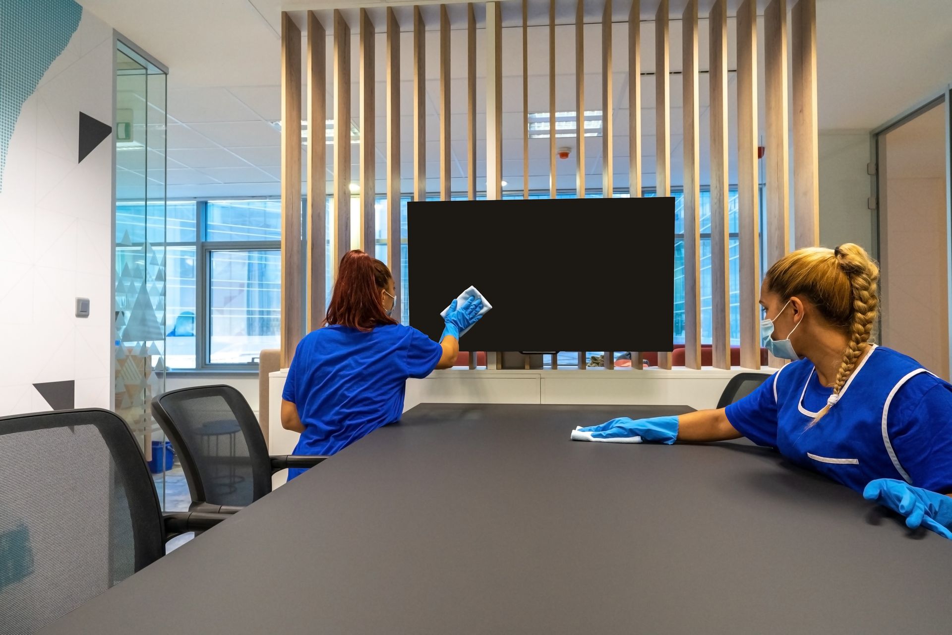 Conference room with two people in blue uniforms cleaning a wall-mounted screen and table