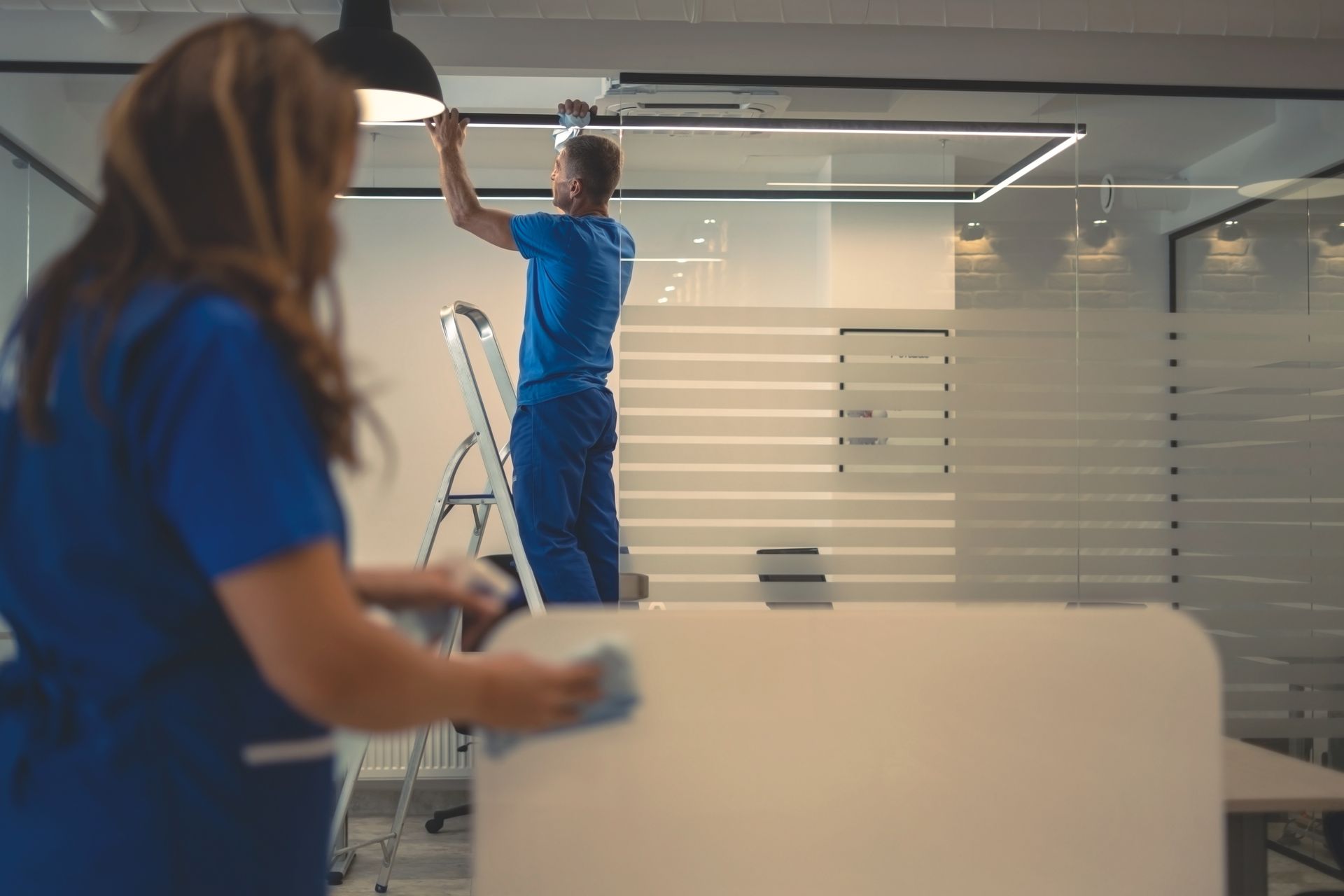 Two workers in blue uniforms installing a ceiling light in a modern office restroom