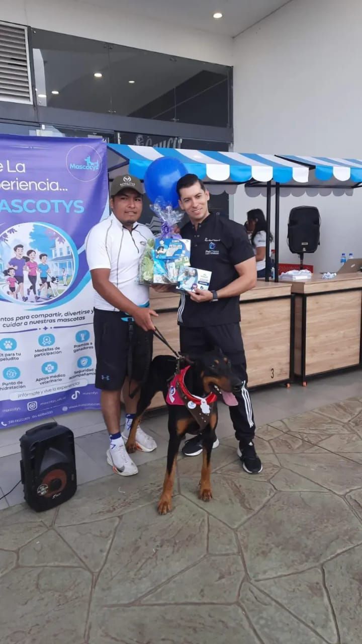 Dos hombres y un dóberman posan con premios en un evento. Una pancarta dice