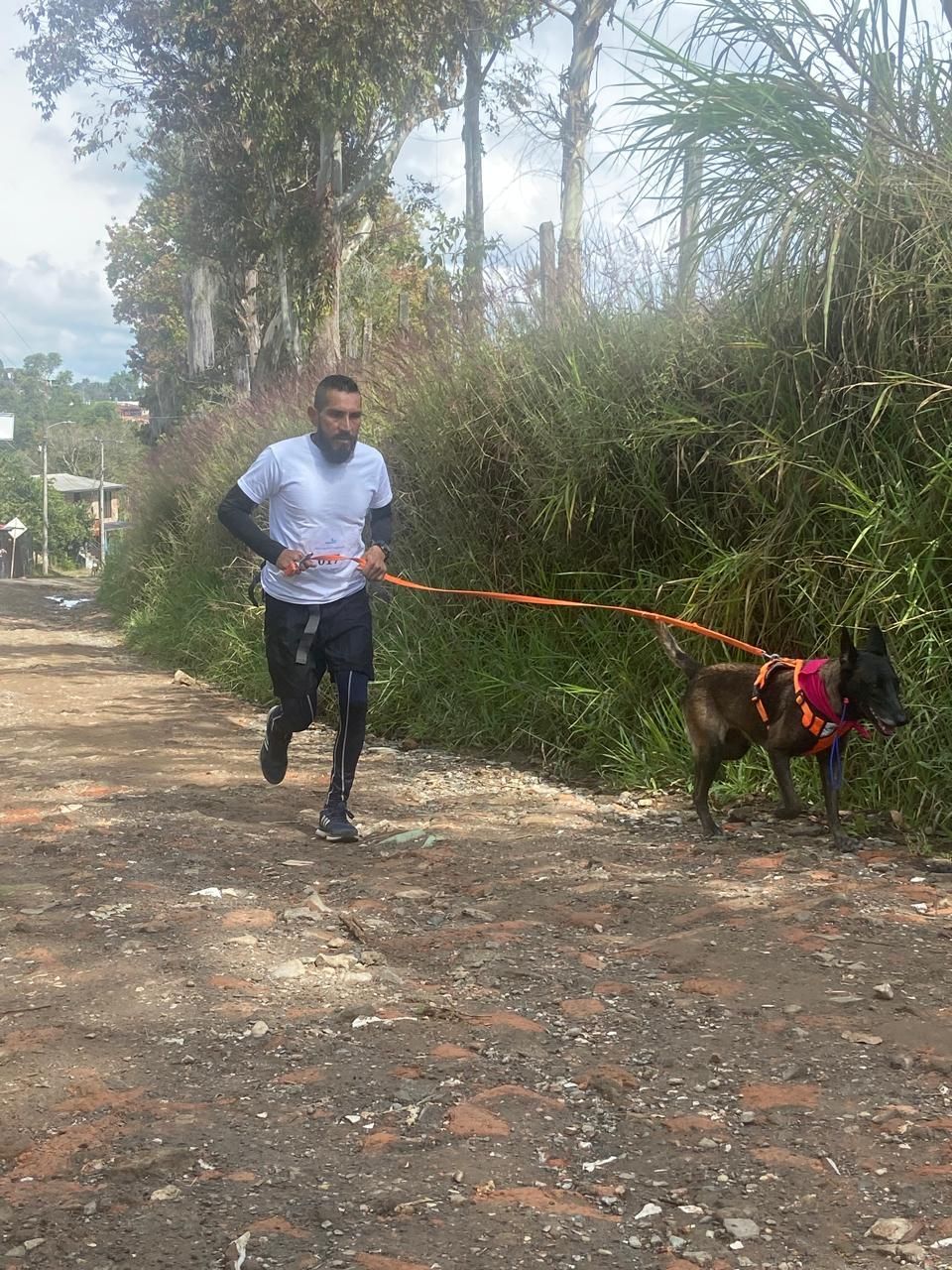Hombre corriendo por un camino de tierra con un perro enganchado a él; árboles y vegetación en el fondo.
