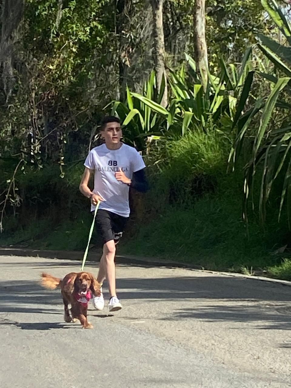 Una persona corriendo con un perro con correa por un camino pavimentado al aire libre, rodeado de vegetación.