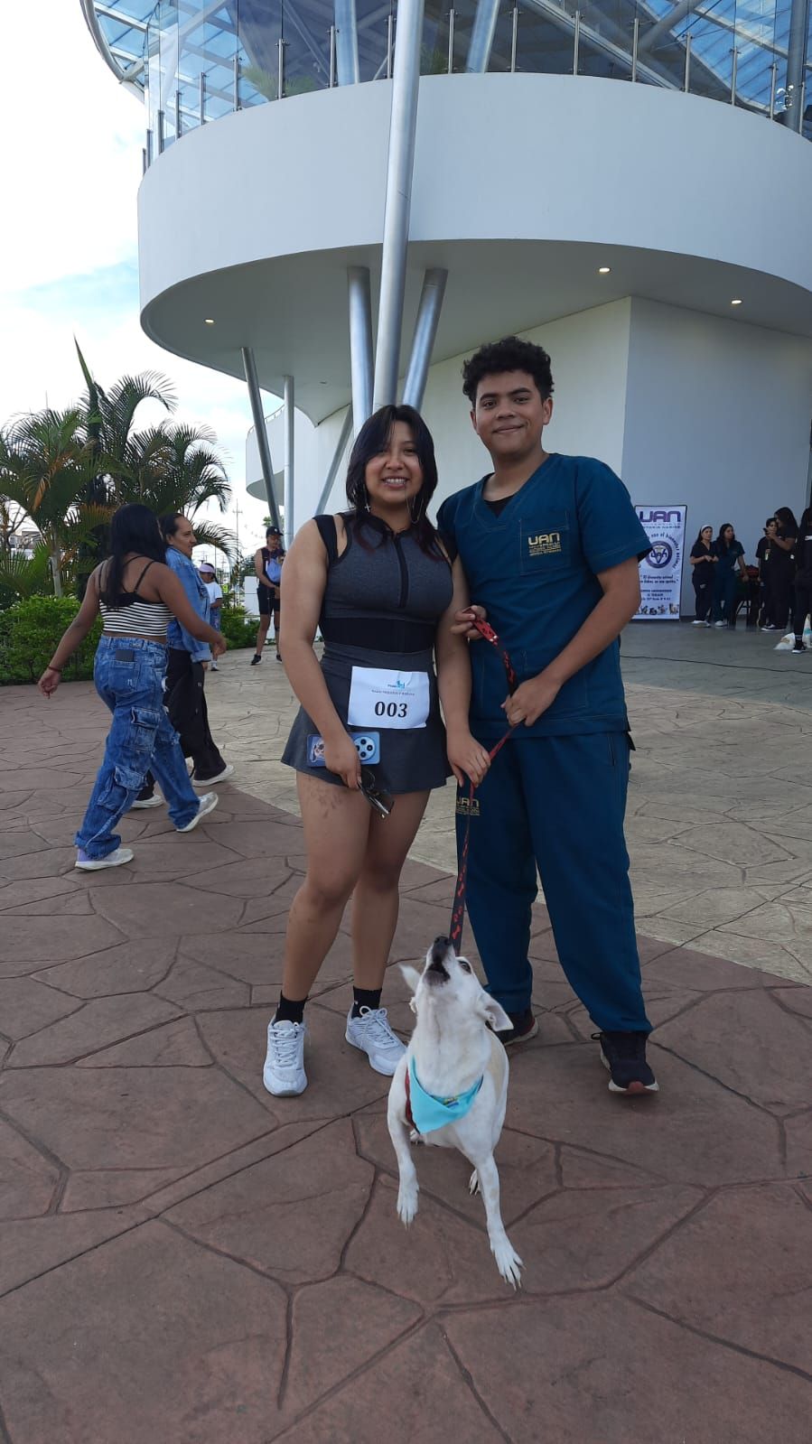 Una mujer y un hombre con un perro en una carrera, posando frente a un edificio. El perro mira hacia arriba.