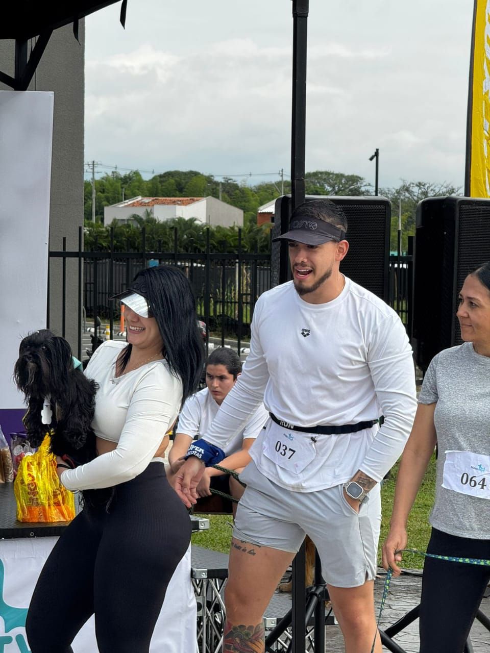 Personas en un evento al aire libre. Una mujer sostiene a su perro, un hombre sonríe y otra mujer está cerca.