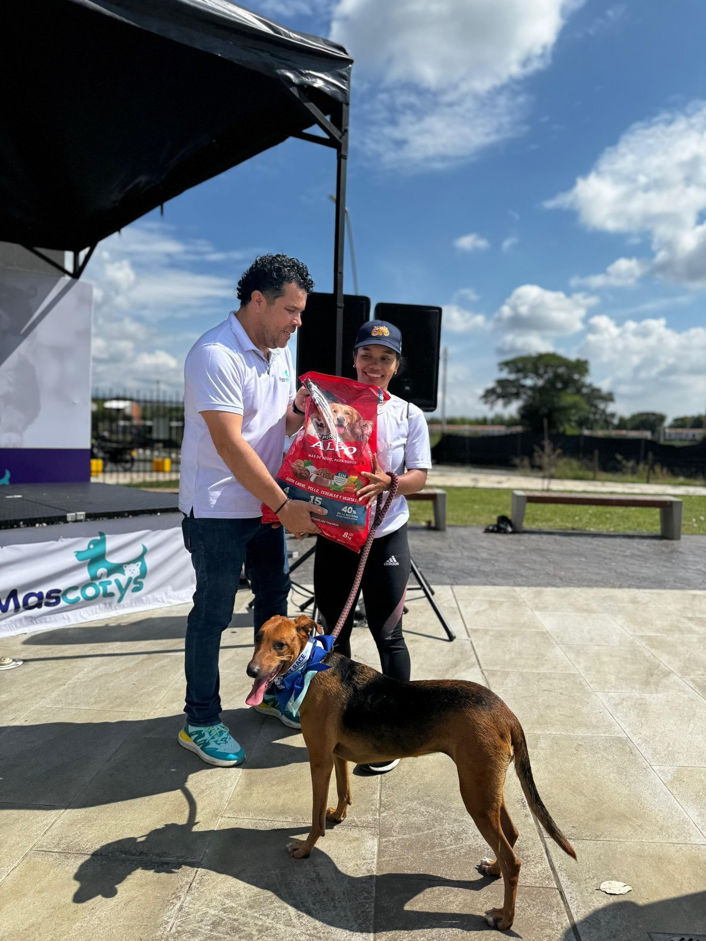 Un hombre le entrega una bolsa de comida para perros a una mujer con un perro con correa en un evento al aire libre.