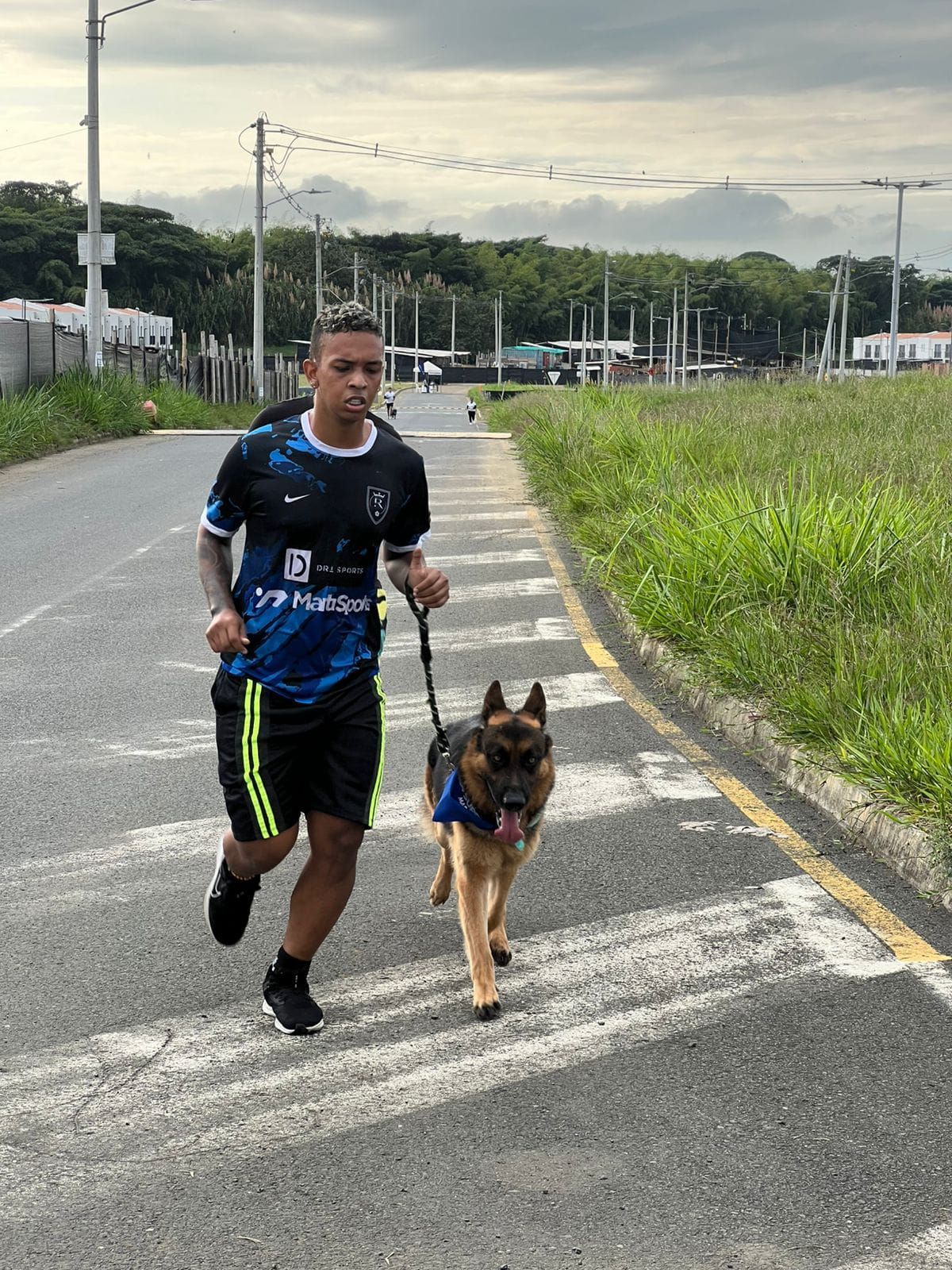 Un hombre trota con un pastor alemán por una carretera asfaltada. Cielo nublado, hierba verde y árboles.