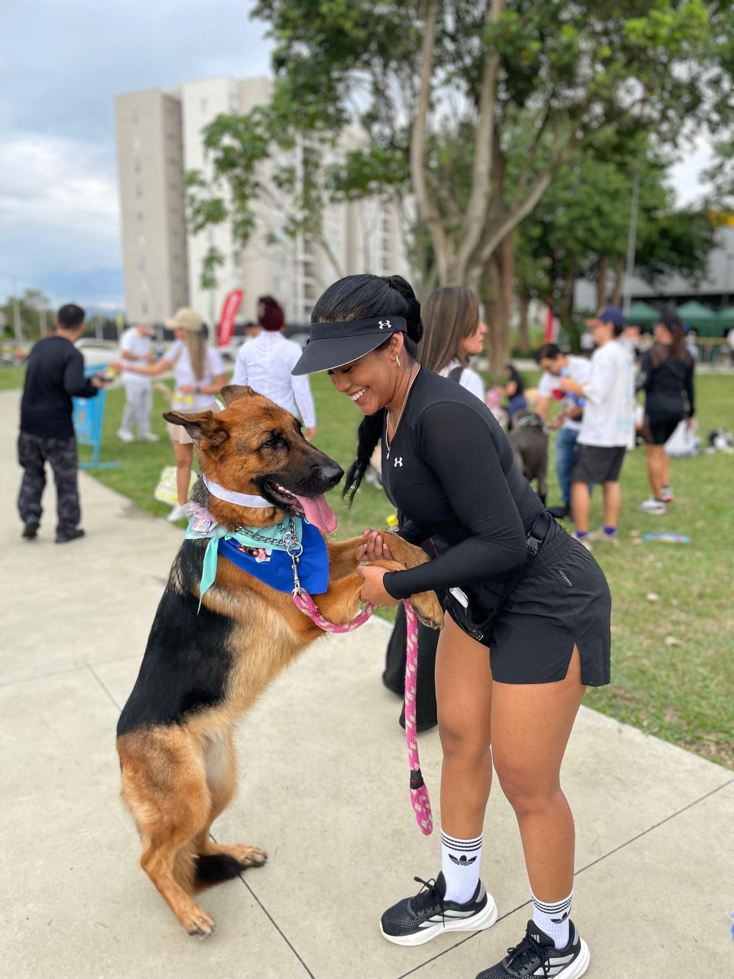 Una mujer con pantalones cortos negros y un sombrero sonríe, sosteniendo las patas de un pastor alemán en un parque.