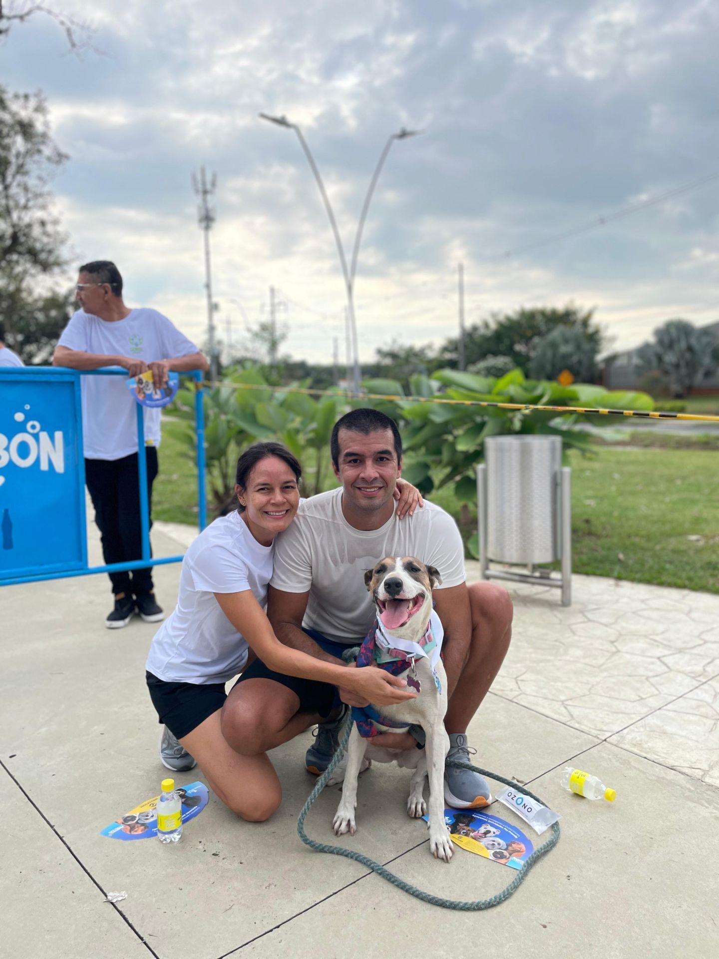 Una pareja con un perro se agacha en un parque. Sonríen, acariciando al perro con un pañuelo azul, cerca de una valla azul.