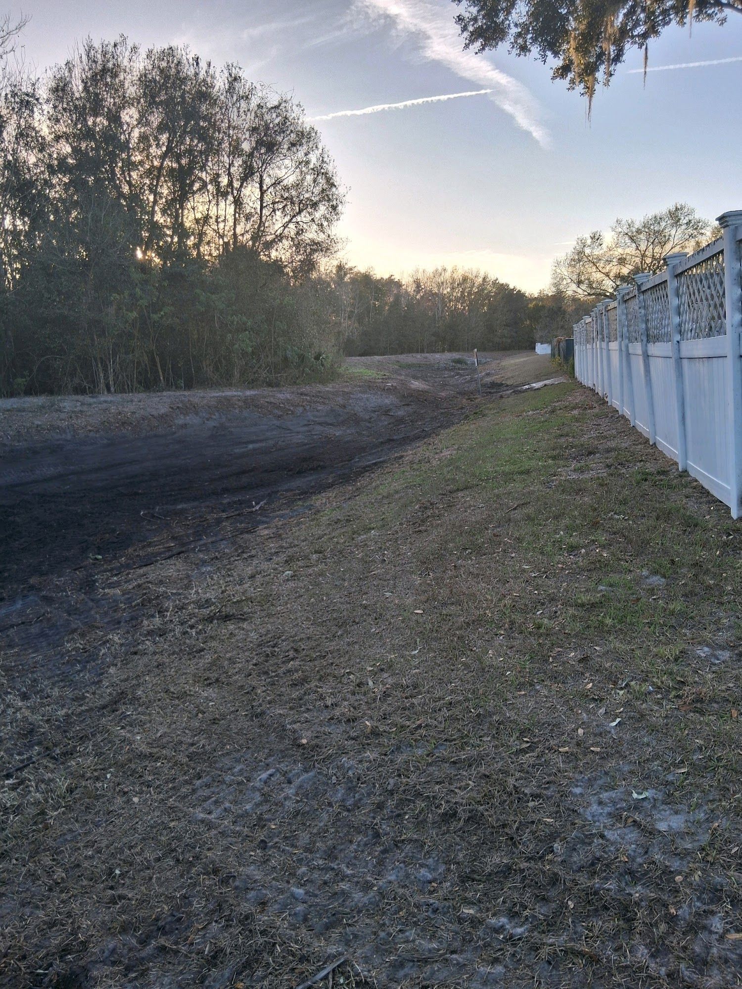 A view of a grassy embankment leading to a dirt patch, beside a white vinyl fence with trees in the background at sunset.