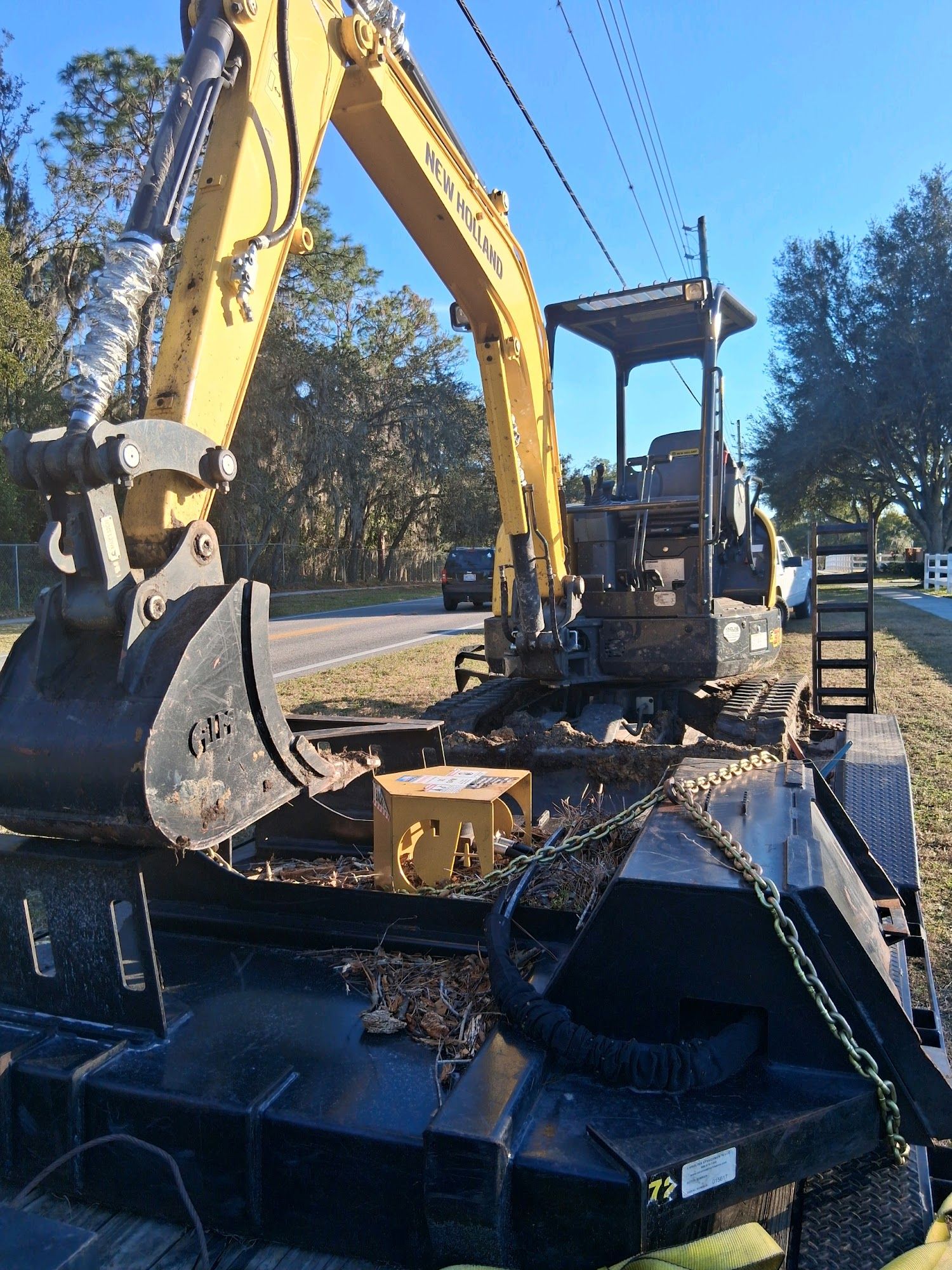 A yellow excavator sits secured on a black flatbed trailer parked on the side of a road on a sunny day.