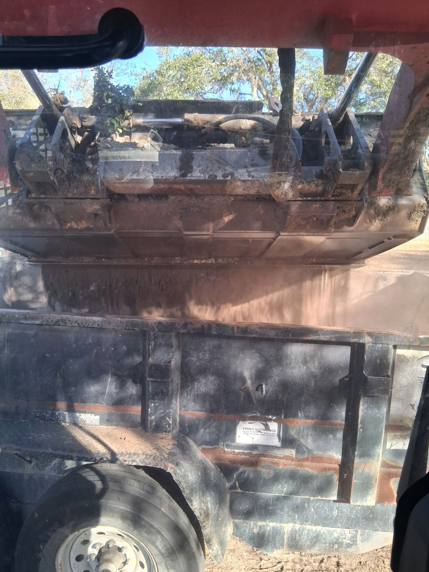A construction vehicle bucket dumps loose dirt into a dark metal trailer in an outdoor, sunlit setting.
