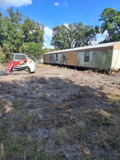 Small red excavator beside a long mobile home in a grassy, wooded lot under blue sky