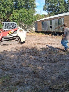 Man on a dirt lot beside a red skid steer and a long white trailer under trees