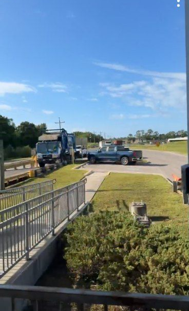 Yellow tow truck and black pickup on a sunny street beside a grassy field and trees.