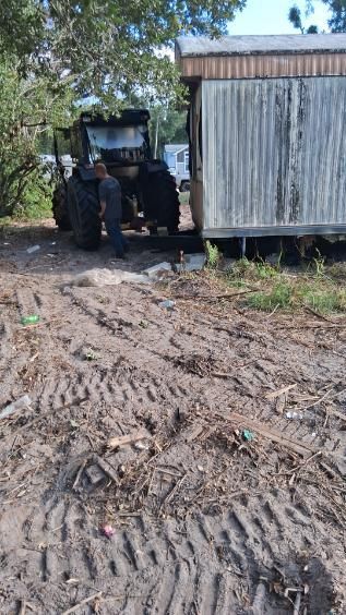 Two people standing by a muddy dirt road beside a small corrugated metal shed and a vehicle under trees.