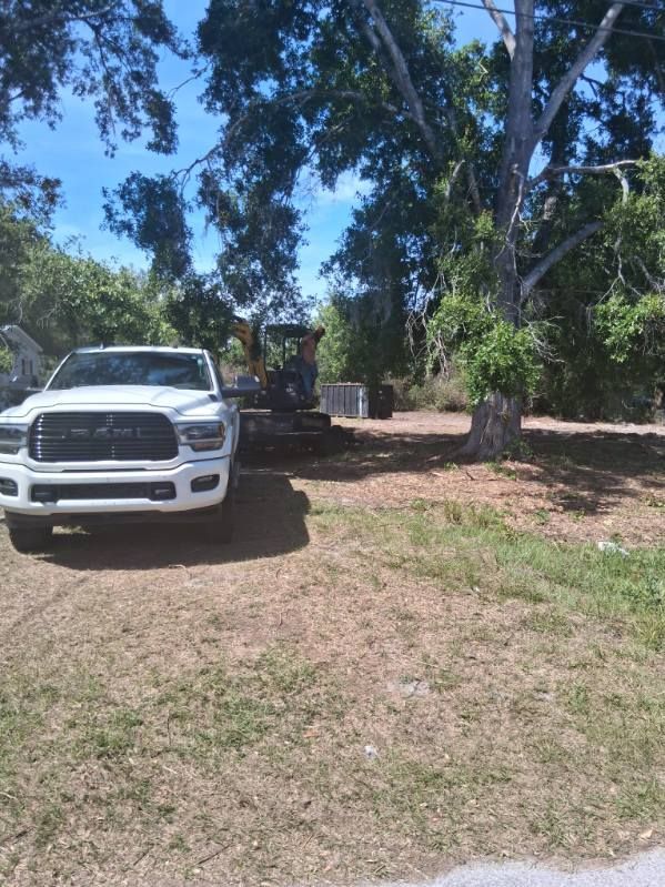 White pickup truck parked under trees in a sunny yard with a flatbed trailer nearby.