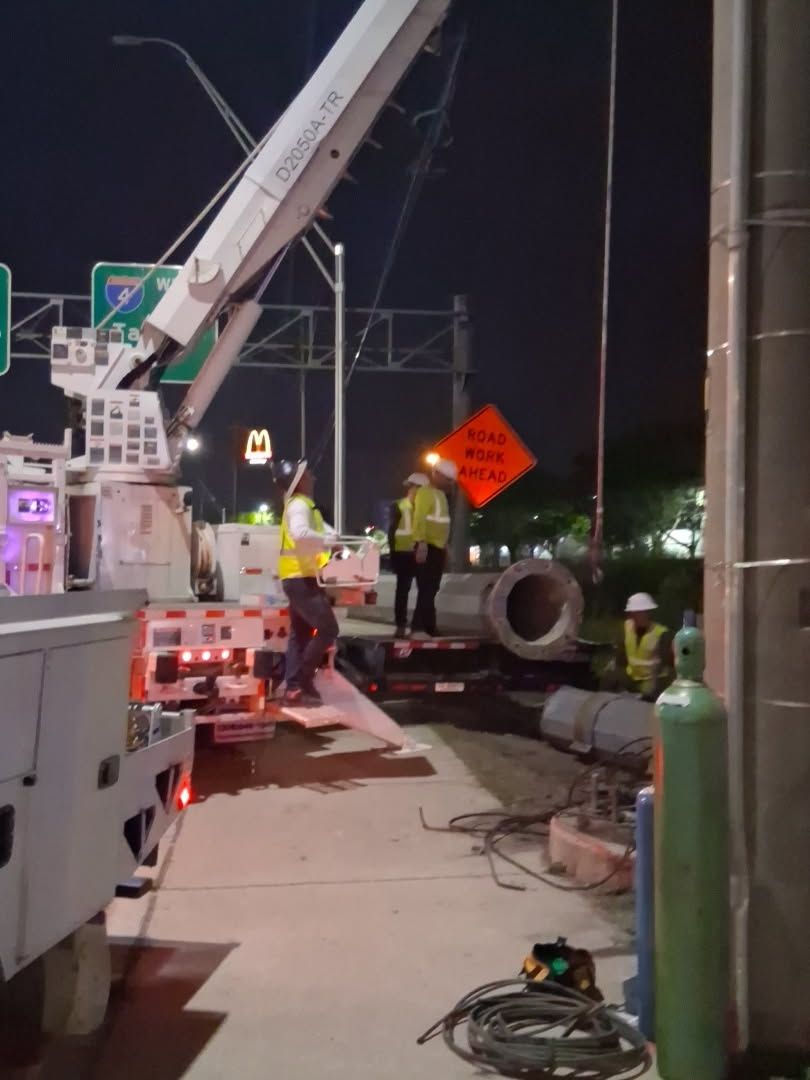 Roadwork at night with a crane, workers in reflective vests, and a large pipe on the sidewalk.