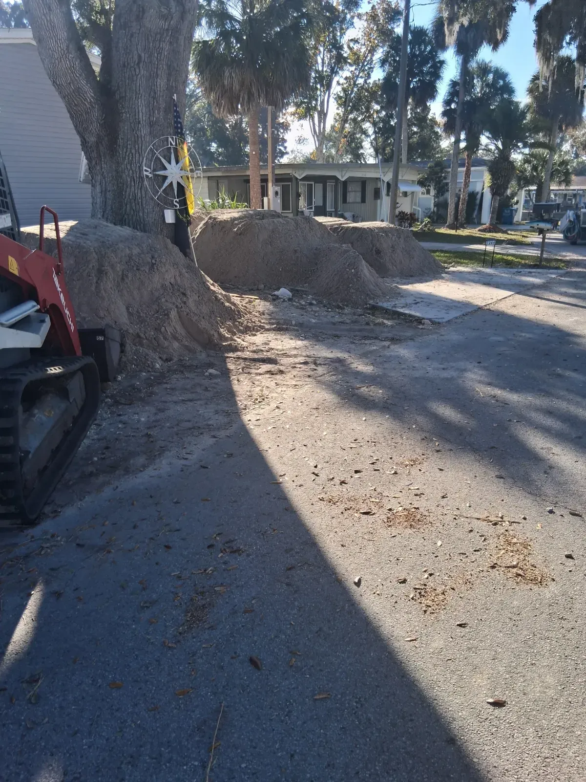 A skid steer loader parked next to piles of dirt beneath a large tree near a residential street.