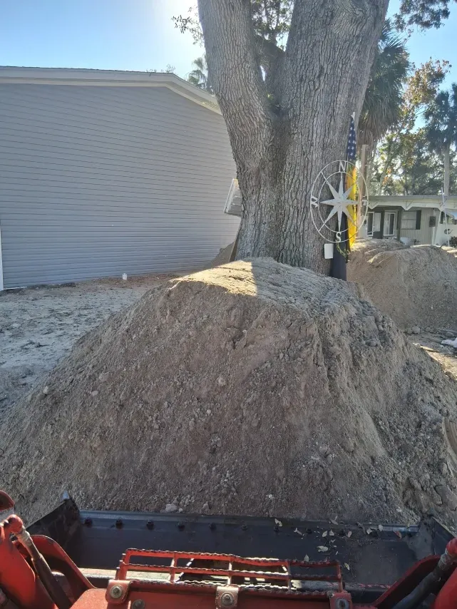 A large pile of dirt sits in front of a tree and a house, viewed from the driver's seat of construction equipment.