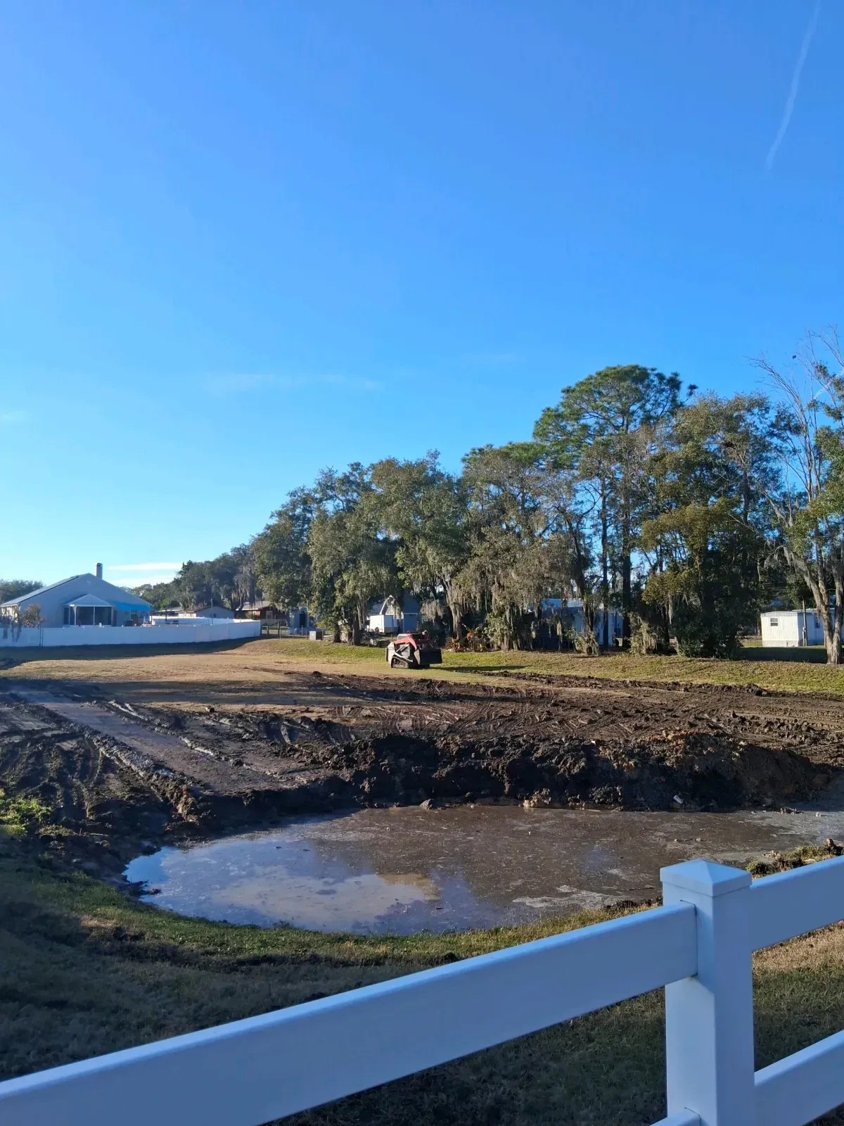 A muddy construction site with a puddle in the foreground, bordered by a white fence under a clear blue sky.