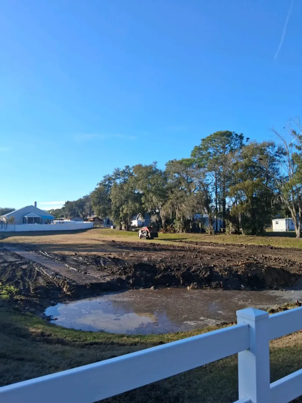 A muddy construction site with a puddle in the foreground, bordered by a white fence under a clear blue sky.